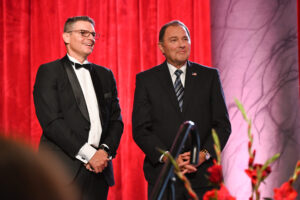 Gary Herbert and Curtis Blair in suits stand on stage at a Utah Valley Chamber of Commerce event.