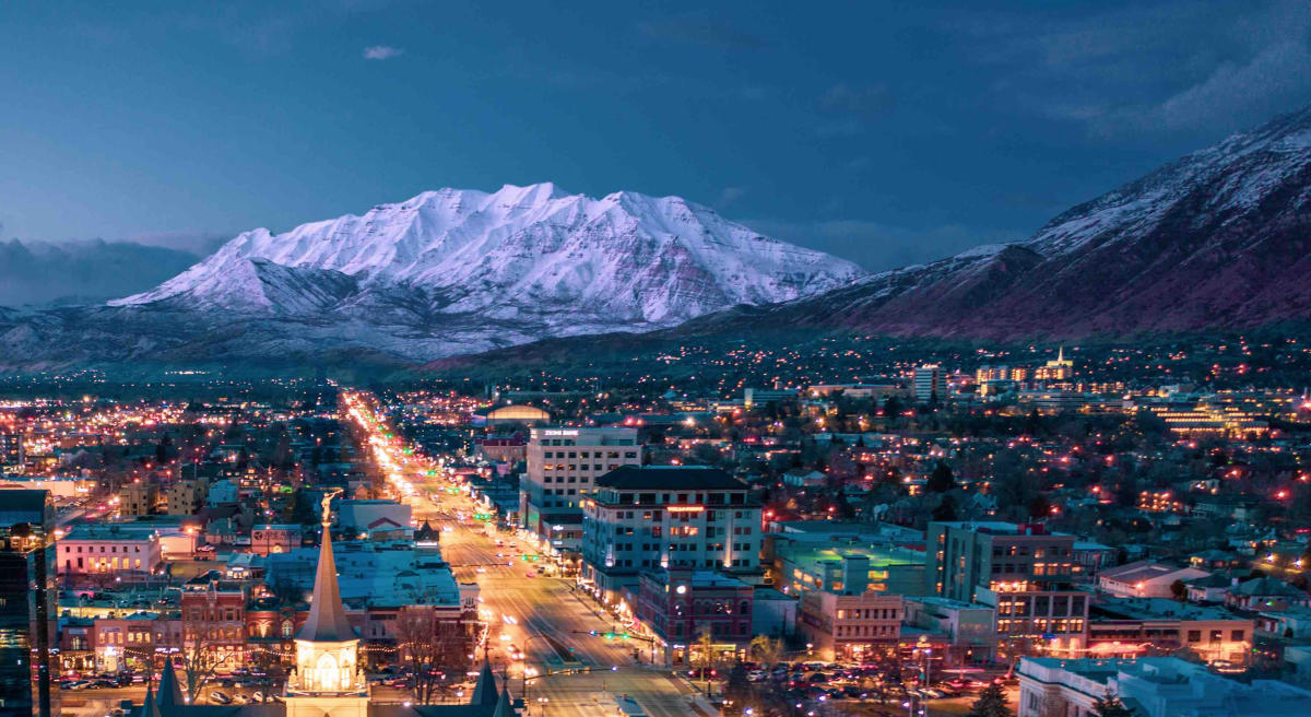 Night view of downtown Provo with city lights and snow-covered mountains in Utah Valley.